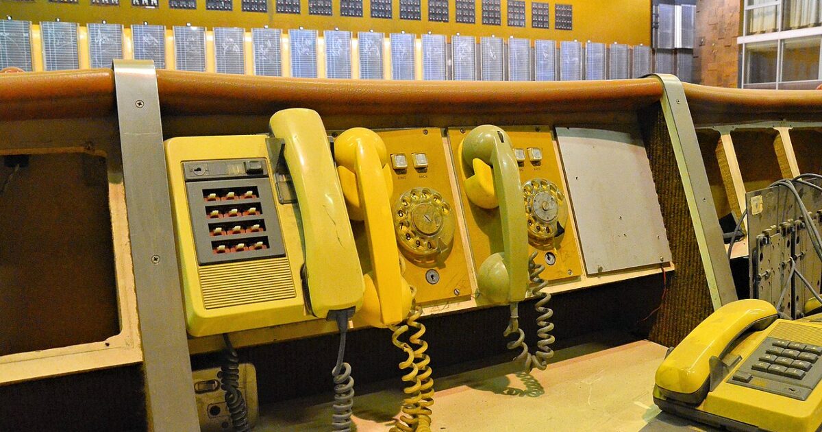 Row of old yellow telephones with coiled cords on a wooden counter in front of a wall of indicator panels