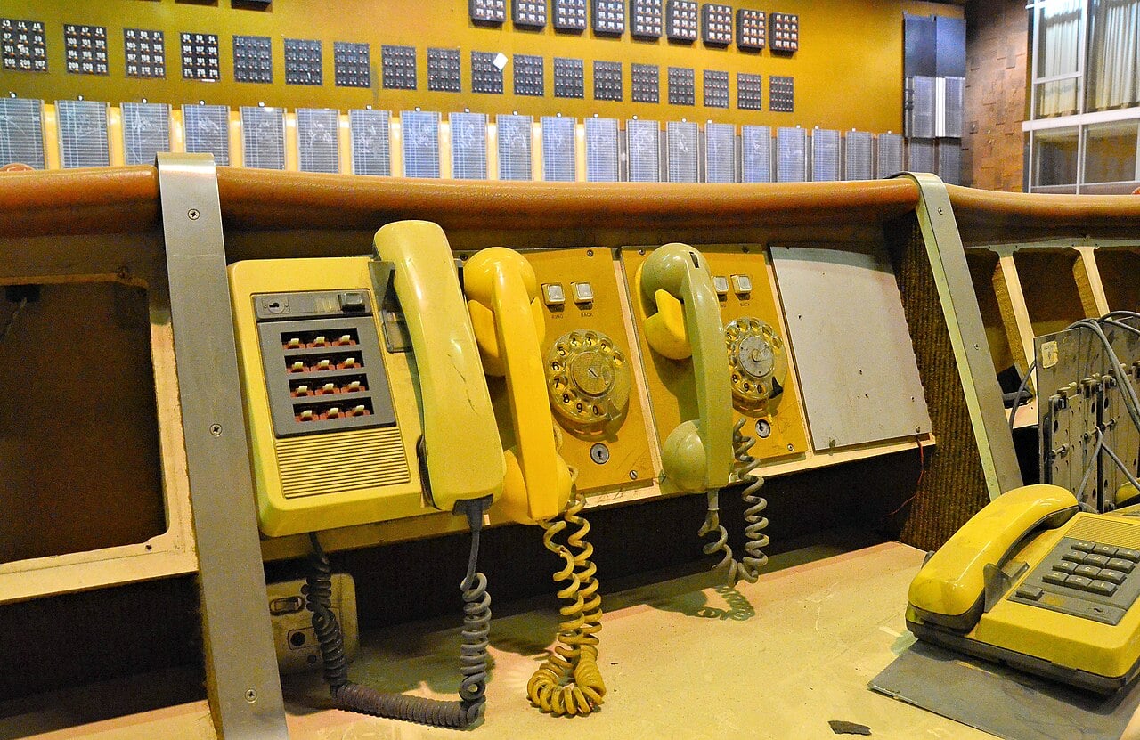 Row of old yellow telephones with coiled cords on a wooden counter in front of a wall of indicator panels