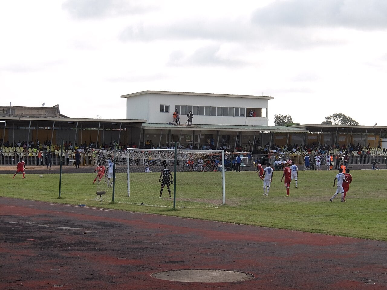 Players in red and white contest a free kick in front of a goal at El Wak Stadium in Accra, with spectators in covered stands and a white press box behind.