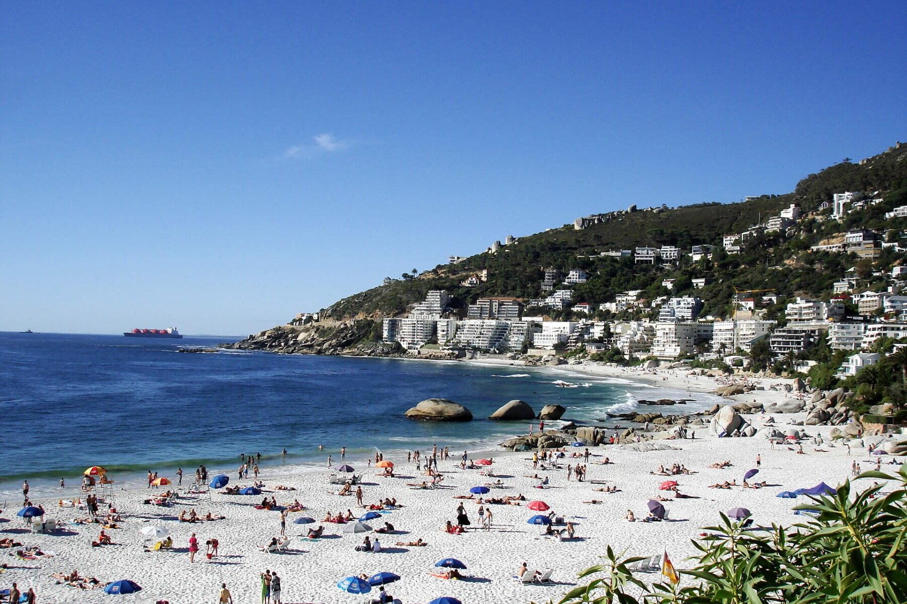 Sunbathers on the white sand of Clifton 4th Beach in Cape Town with apartments on the hillside and a ship on the horizon