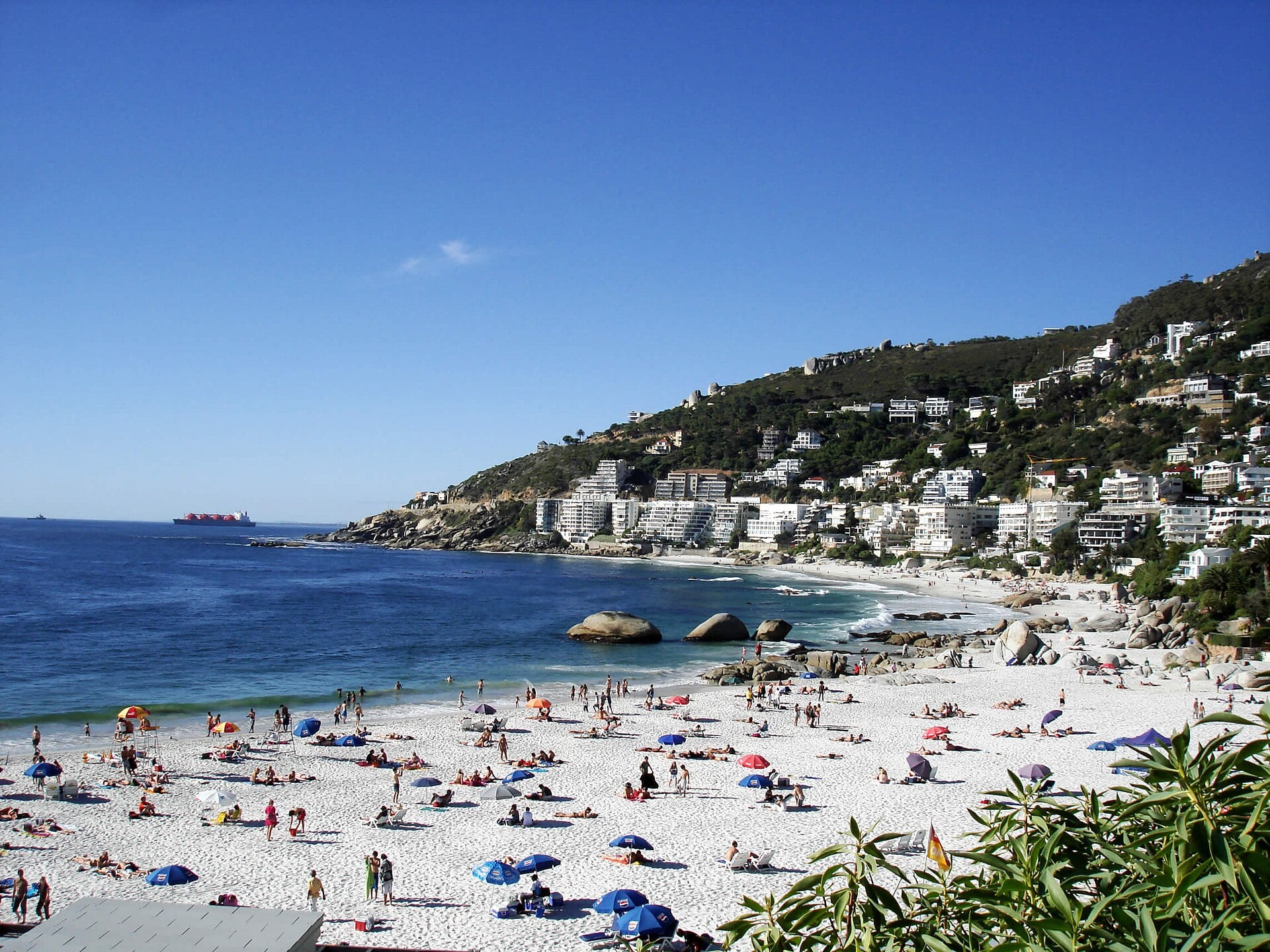Sunbathers on the white sand of Clifton 4th Beach in Cape Town with apartments on the hillside and a ship on the horizon