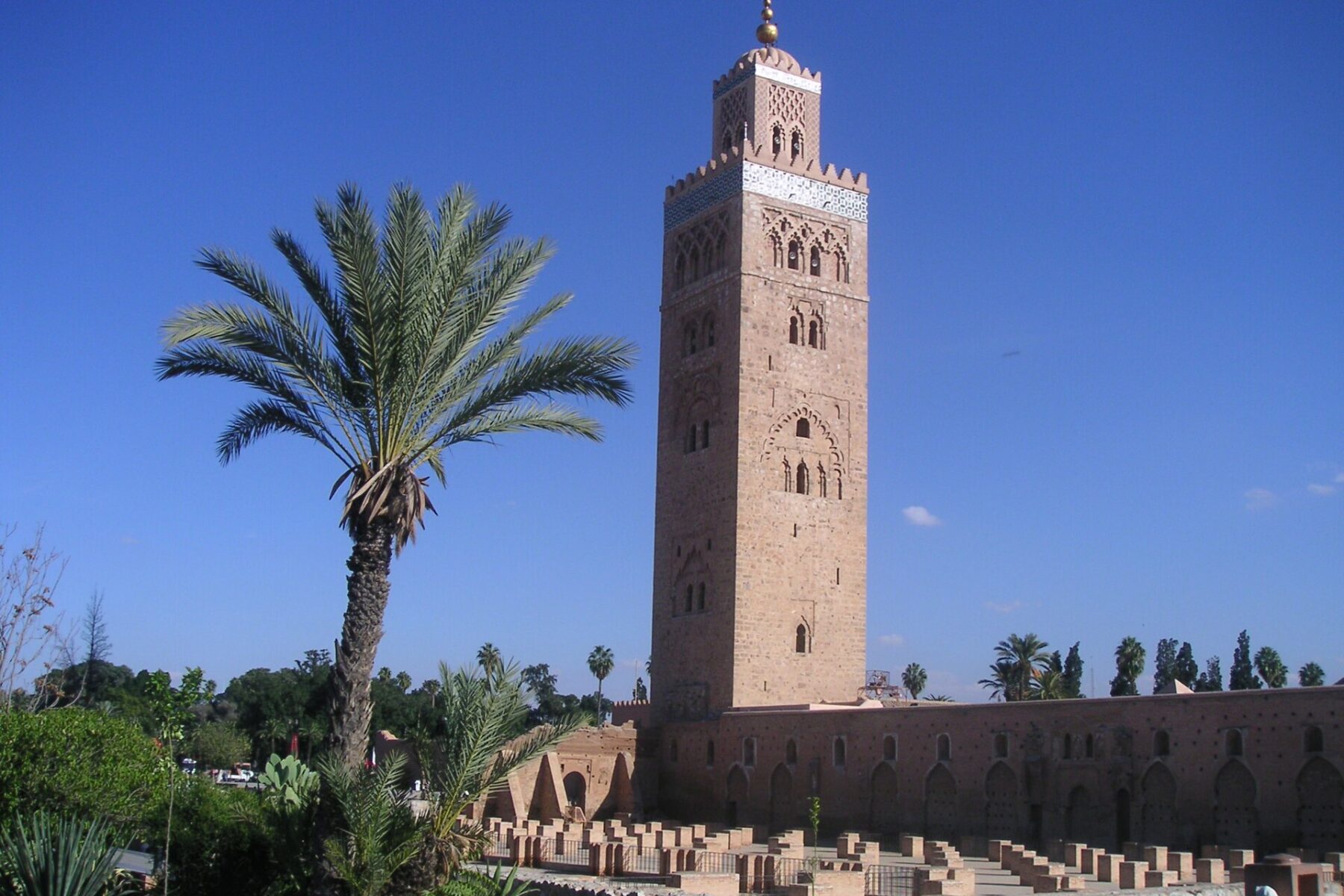 The Koutoubia minaret in Marrakech rising above a palm tree and the low ruined walls beside the mosque under a deep blue sky