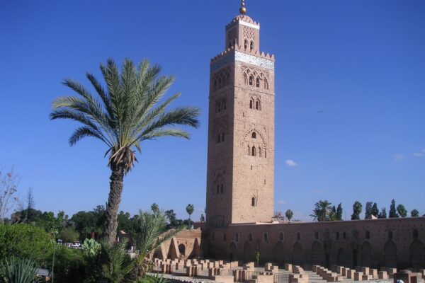 The Koutoubia minaret in Marrakech rising above a palm tree and the low ruined walls beside the mosque under a deep blue sky