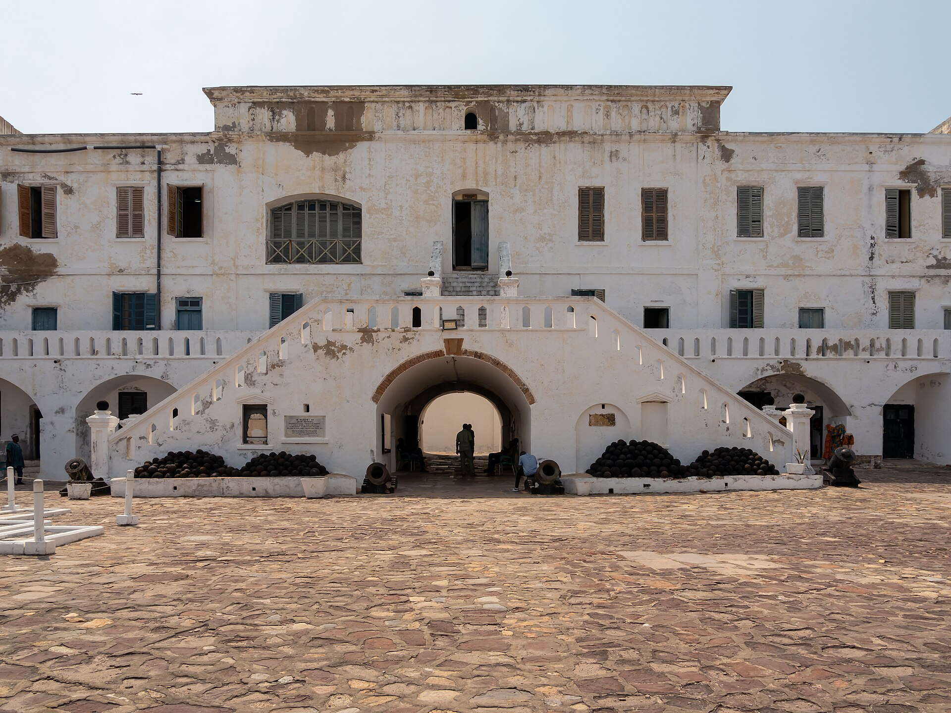 White courtyard facade of Cape Coast Castle with a double staircase, an arched passage and piles of cannonballs on a cobbled yard