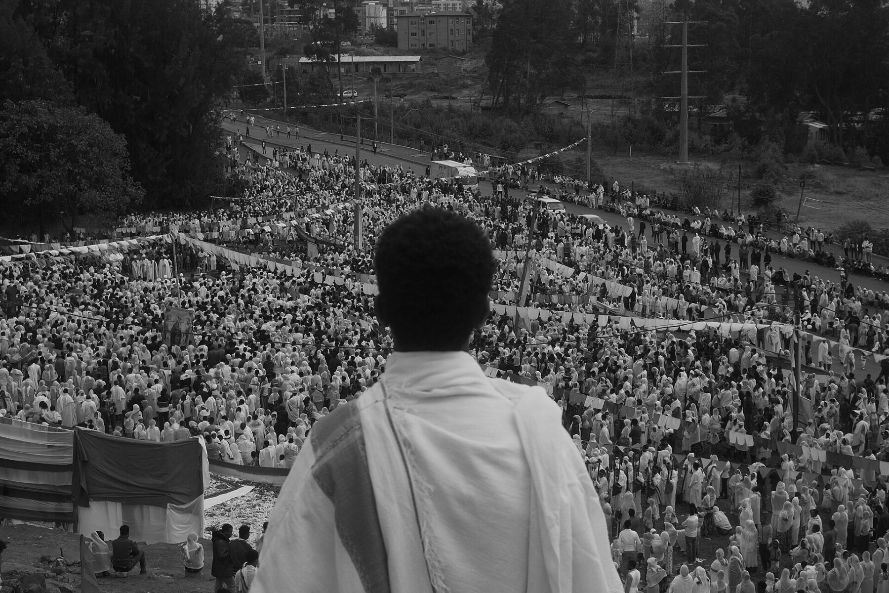 Black-and-white view over a large crowd of worshippers in white shawls at a Timkat celebration in Ethiopia, seen from behind a standing man