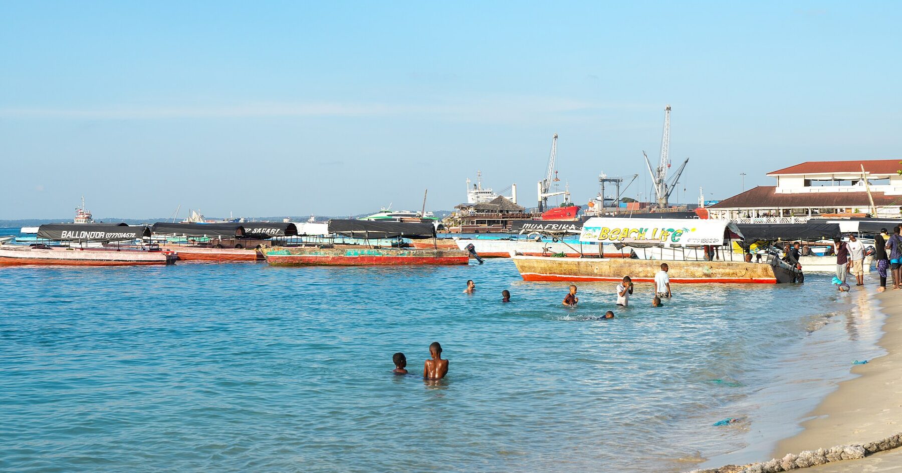 Wooden boats moored in shallow turquoise water off a sandy beach, with children swimming and port cranes in the background