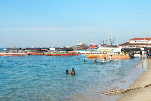 Wooden boats moored in shallow turquoise water off a sandy beach, with children swimming and port cranes in the background