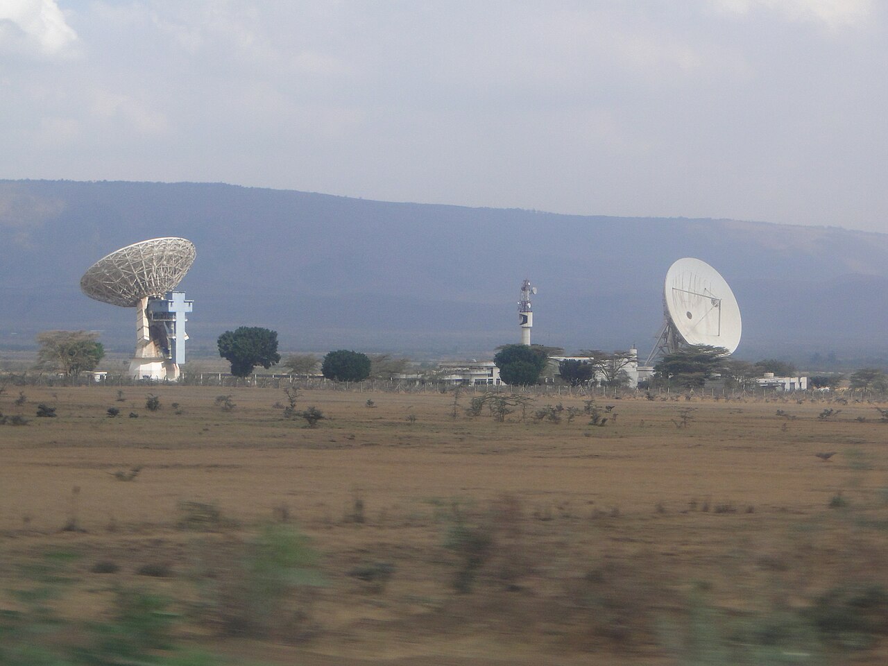 Two large white satellite dishes and a radio tower on dry grassland with hills behind, at Longonot in Kenya