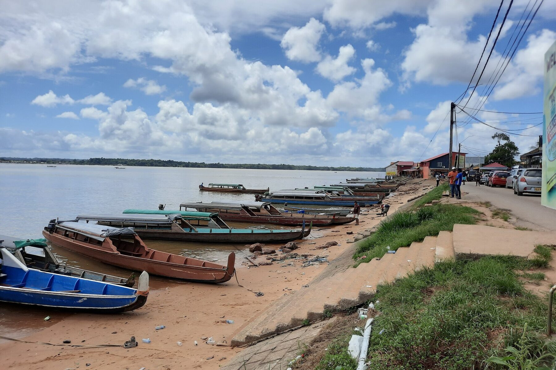Long wooden passenger boats pulled up on the sandy bank of the Marowijne river at Albina, with shops, parked cars and a street along the shore and the French Guiana bank on the far side