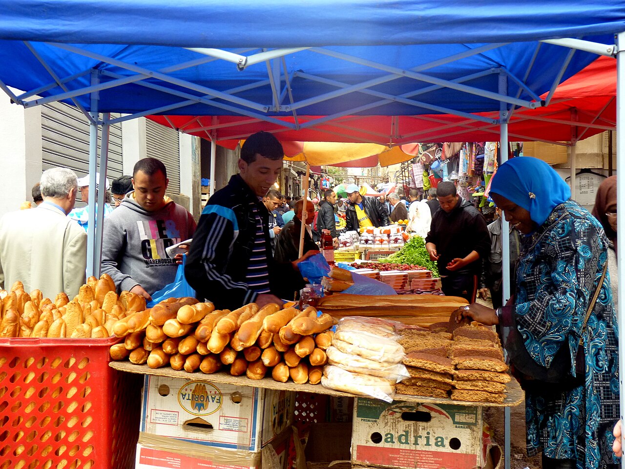 Shoppers and sellers at a bread and cake stall under blue and red canopies in an Algerian street market