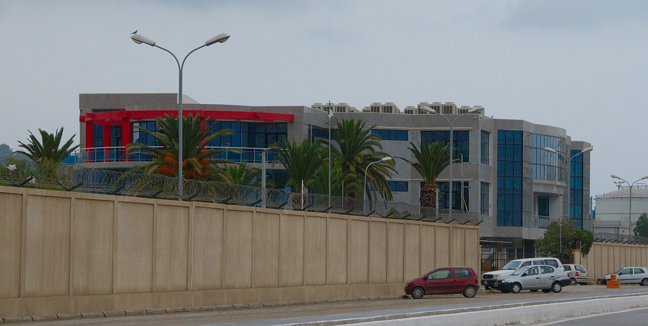 A modern glass-fronted Sonatrach office building behind a perimeter wall in Béjaïa, Algeria