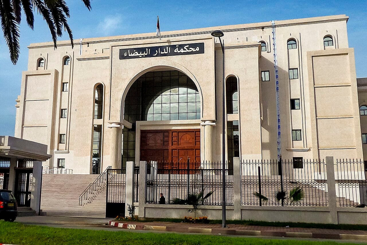 A cream-coloured court building with a tall arched glass entrance, wooden doors and the Arabic sign of the Dar El Beida court, behind an iron fence