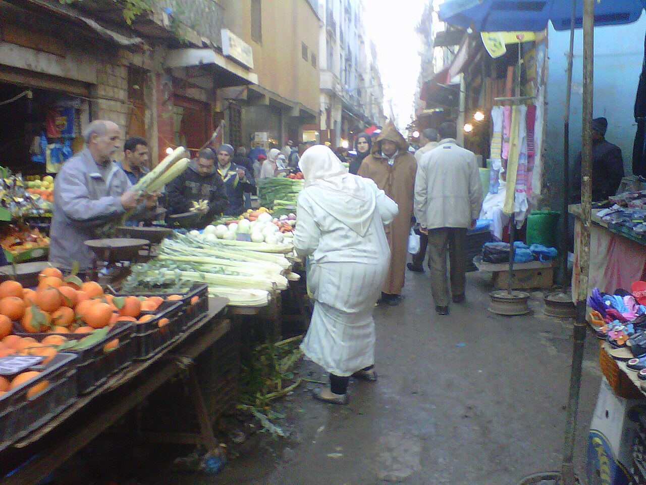 A shopper in a white robe walks past fruit and vegetable stalls in a crowded market street in Oran, Algeria