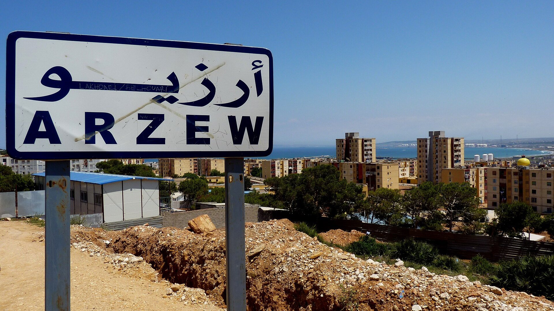 Road sign for Arzew with apartment blocks and the Mediterranean coast behind