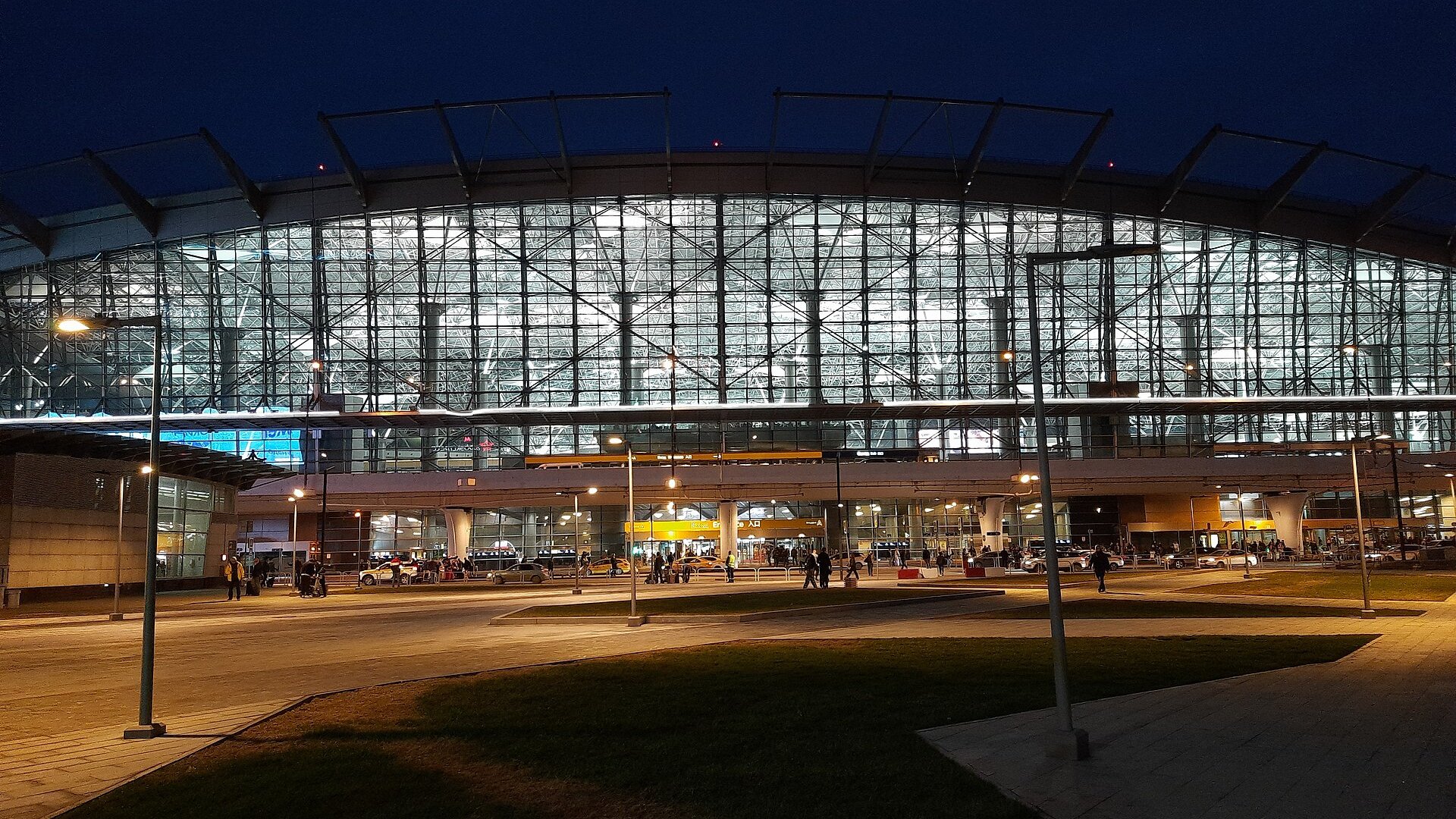 The glass front of the passenger terminal at Vnukovo International Airport in Moscow, lit at dusk with cars and travellers outside