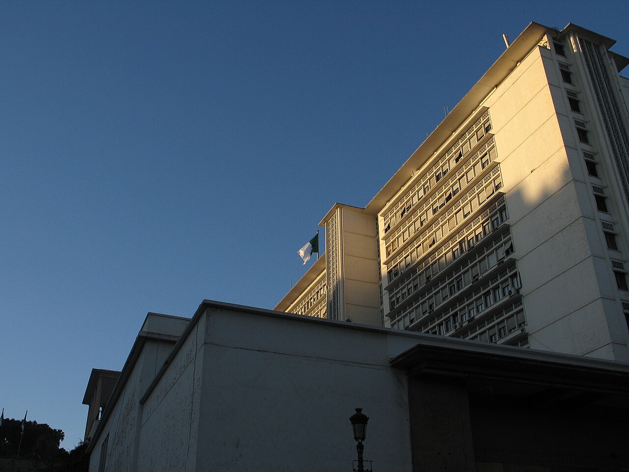 The tall, pale government tower in Algiers, with the Algerian flag on the roof, seen from below at dusk