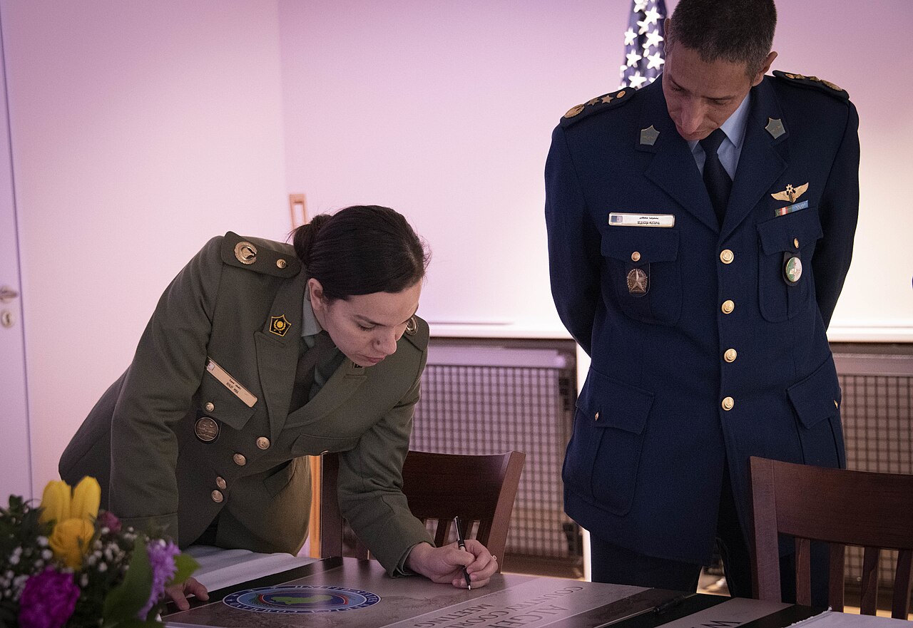 Two Algerian army officers in dress uniform sign meeting minutes at a table, one bending over the page and one standing beside her