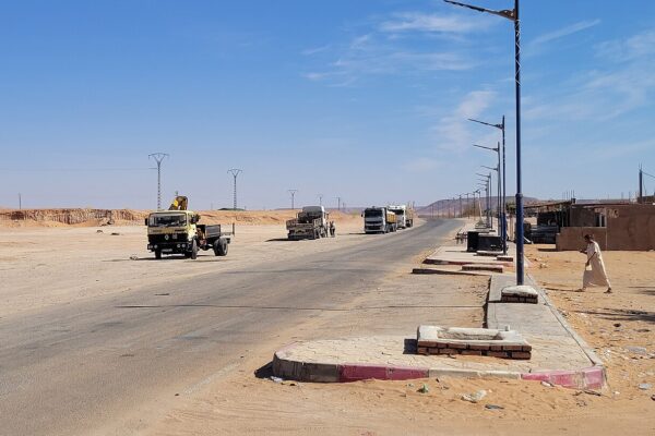 Heavy trucks parked along a desert highway in southern Algeria, with a yellow crane truck in the foreground and power lines behind