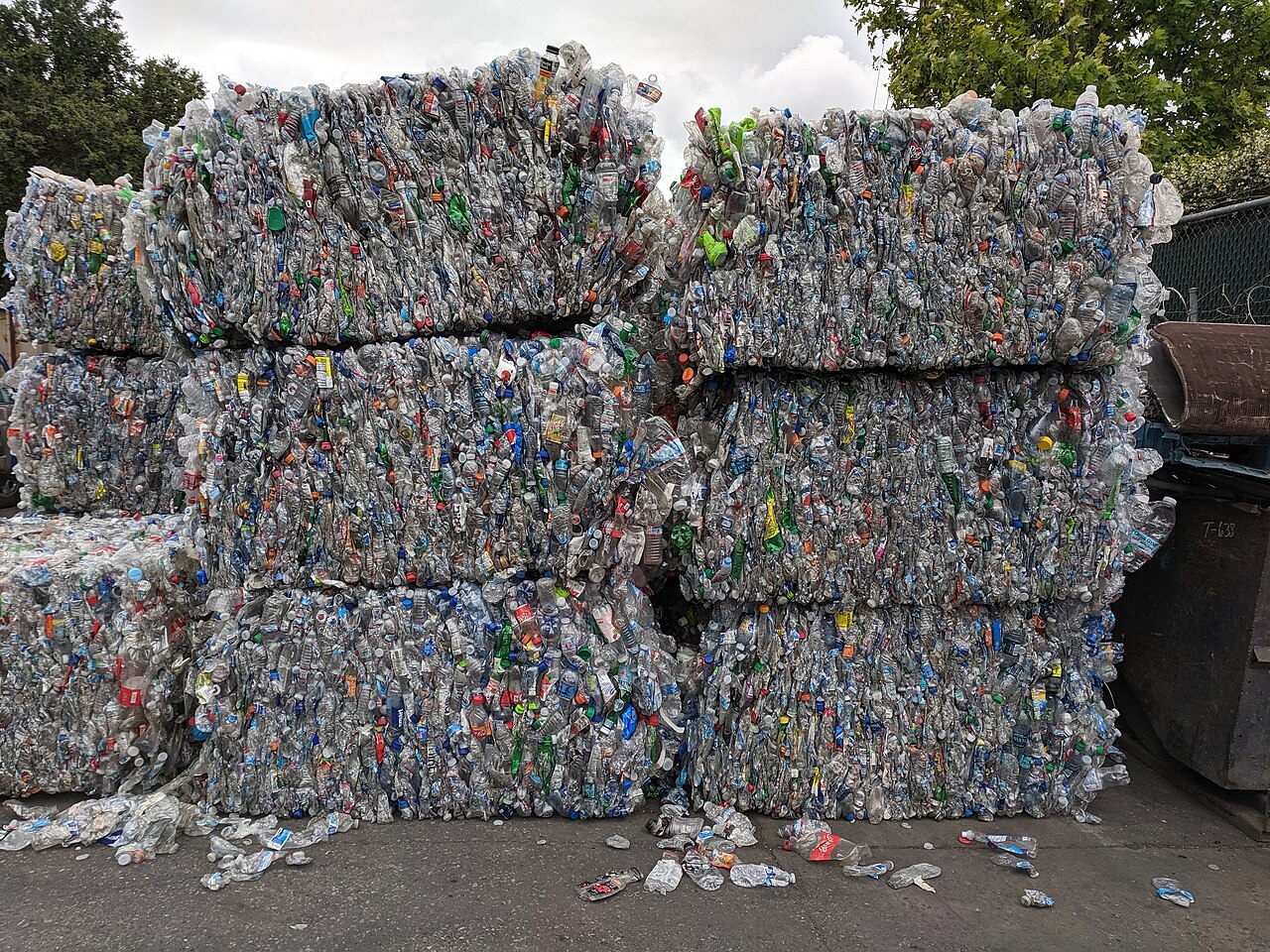 Stacked bales of crushed PET plastic drink bottles at a recycling yard, with a fence and trees behind