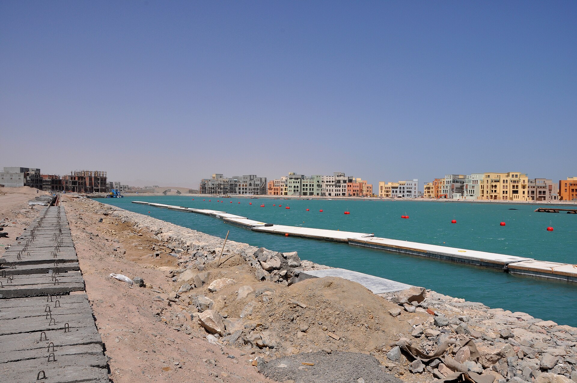 Turquoise marina channel with floating pontoons and red buoys, lined by pastel-coloured apartment blocks in El Gouna, with a stony shore in the foreground