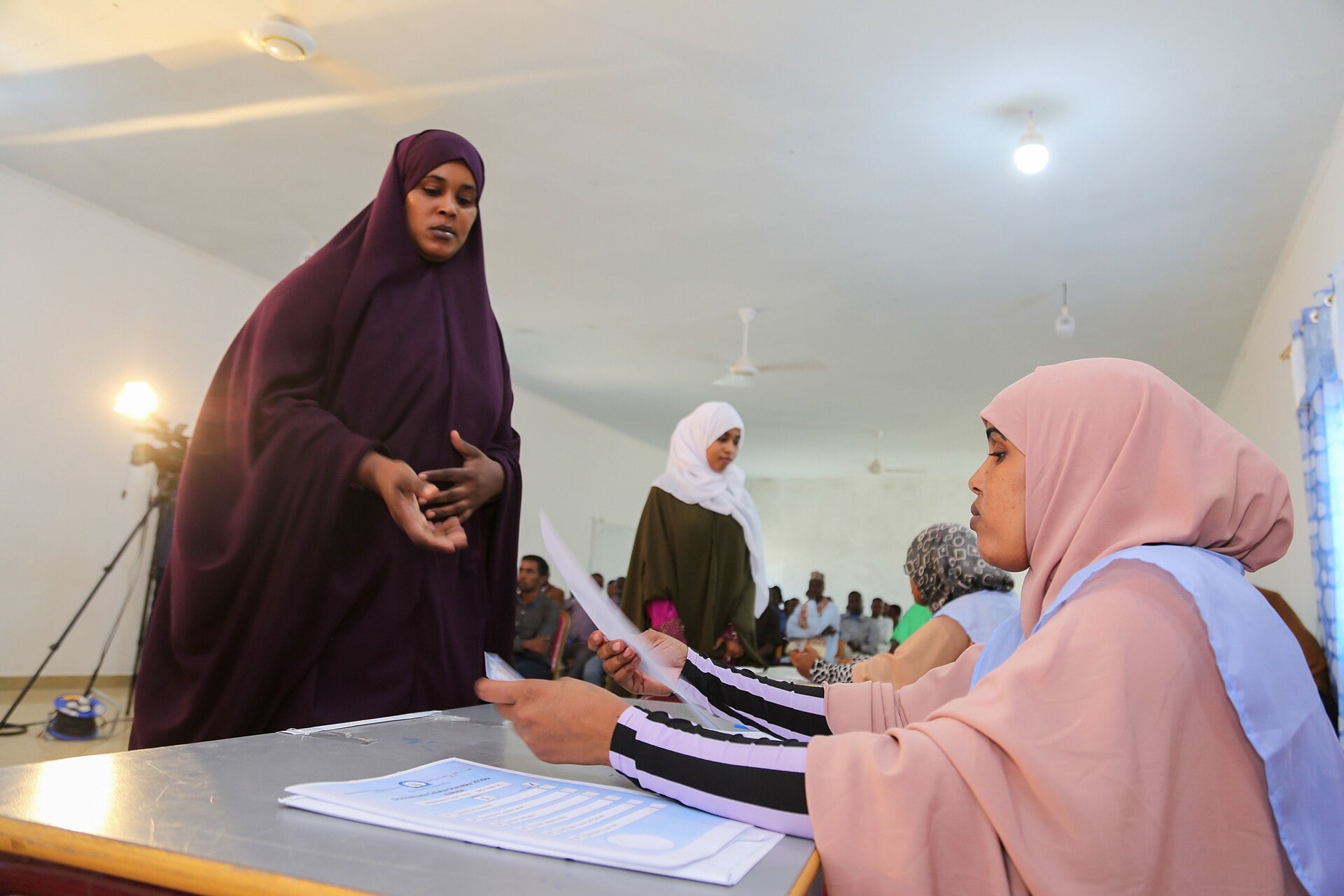 An electoral official in a pink headscarf hands a ballot paper to a woman delegate in a dark purple dress at a table in a hall