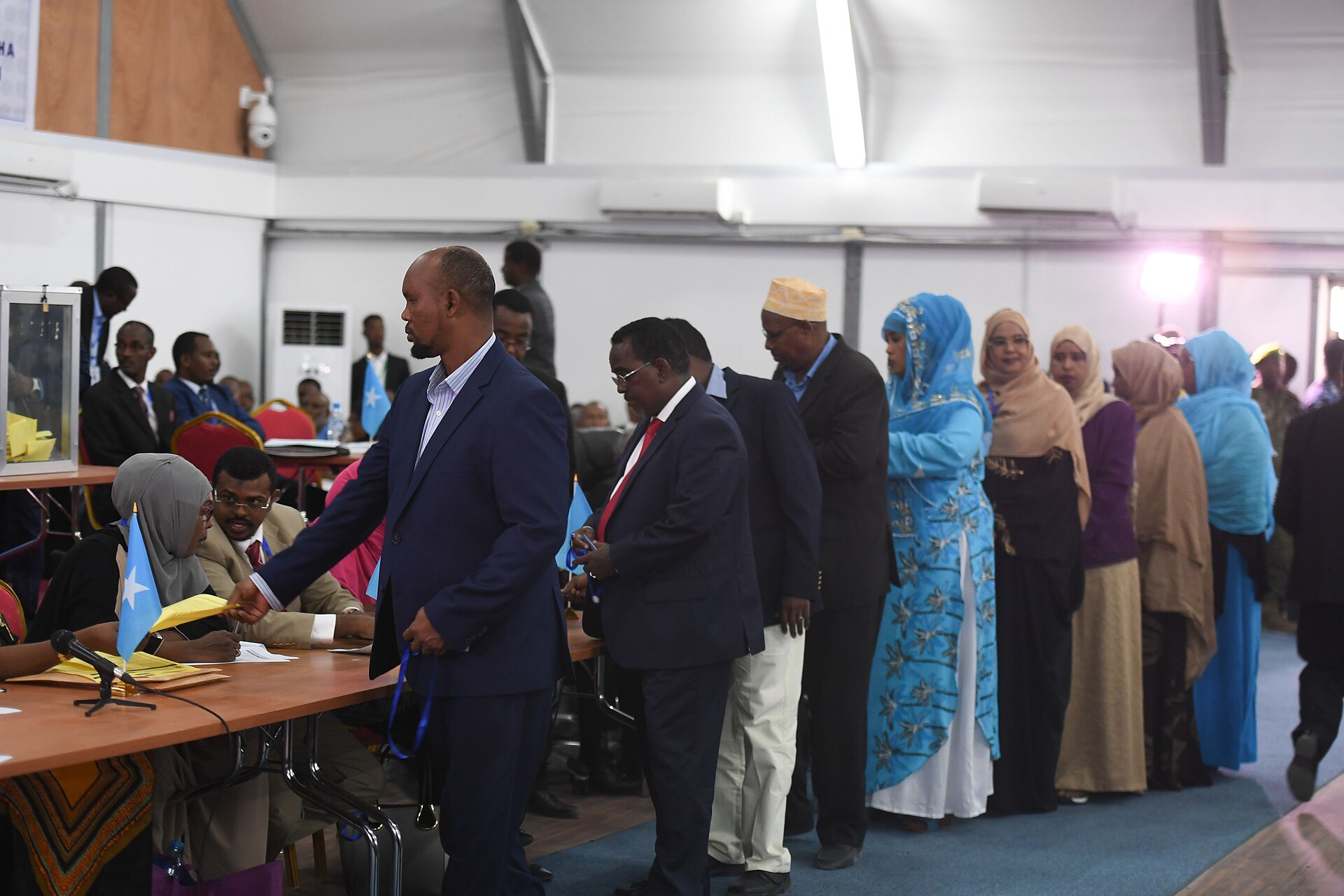 A line of men and women in suits and colourful dresses queue beside registration tables in a hall with Somali flags