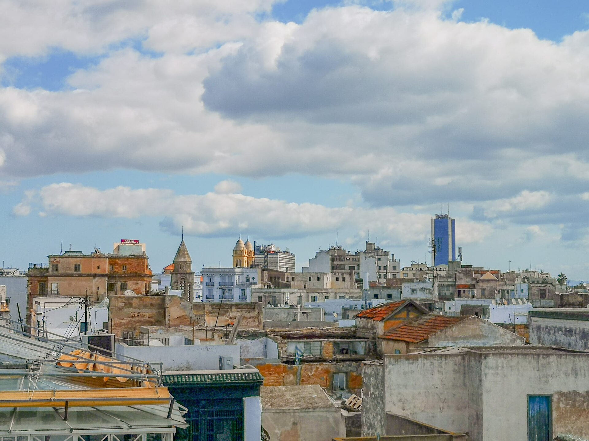 Rooftops of the old city of Tunis with a church tower, a domed building and a blue office tower under a cloudy sky