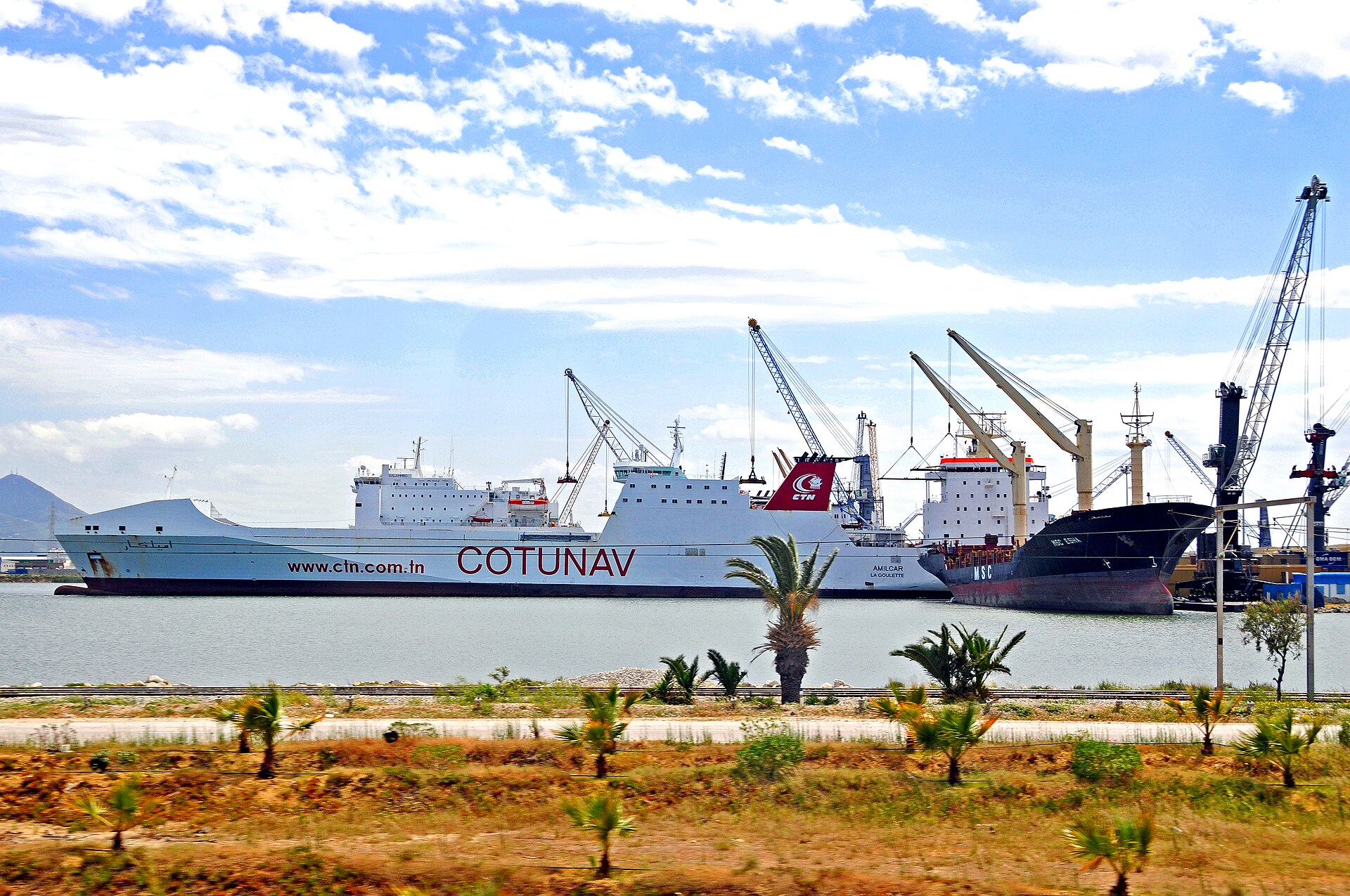 A white Cotunav ferry and a dark cargo ship moored at a quay with cranes under a blue sky with clouds
