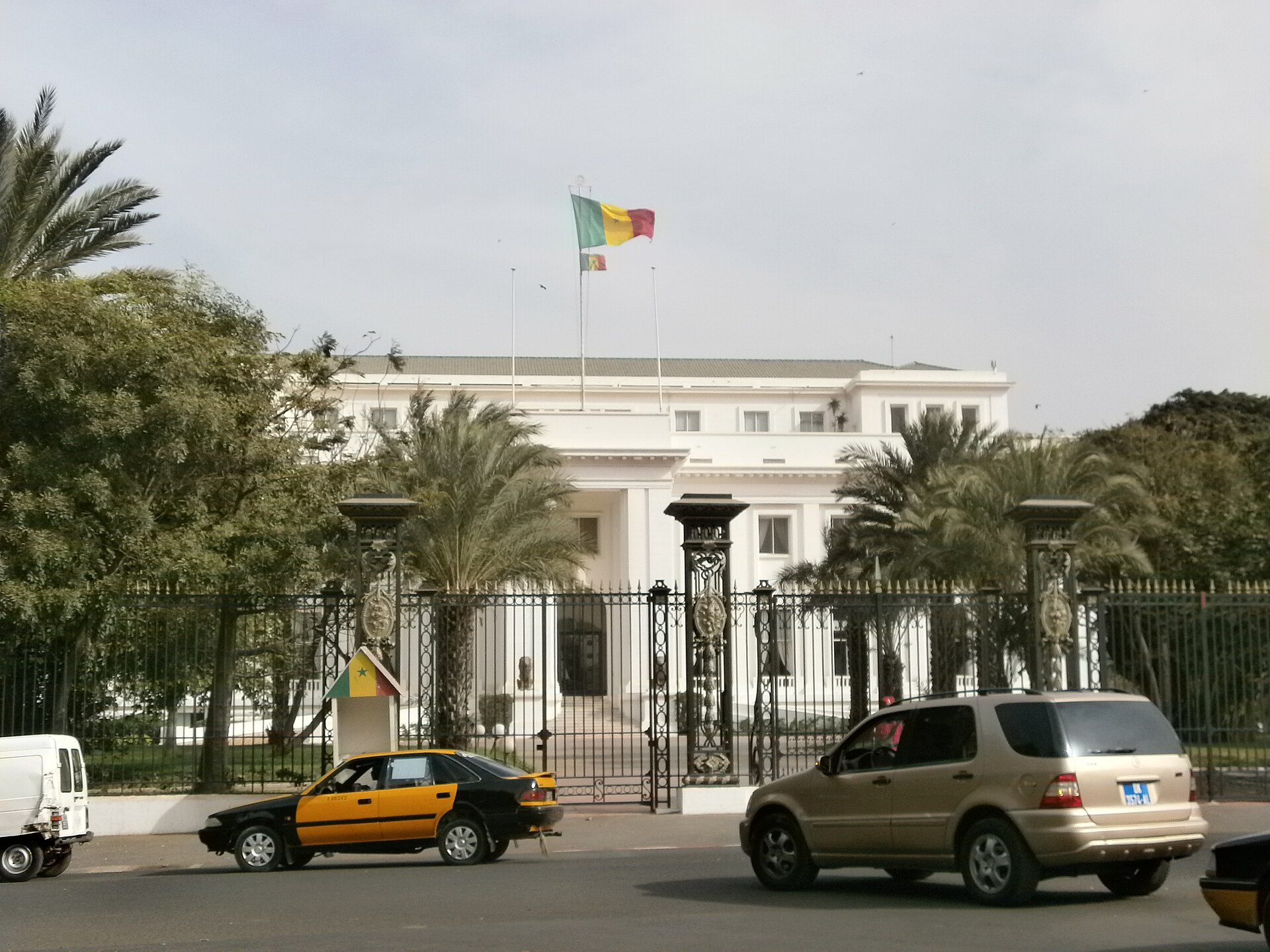 White presidential palace in Dakar behind an iron fence, with the Senegalese flag flying, palm trees, a taxi and a car in the street
