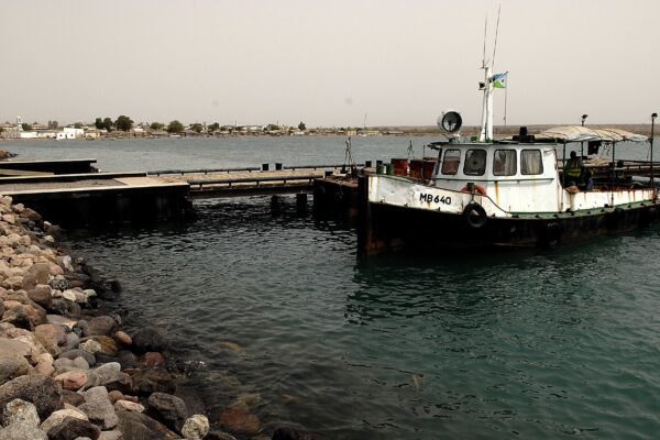 A white and black tugboat flying the Djibouti flag moored at a pier in Obock harbour, with rocks in the foreground and the low town on the far shore