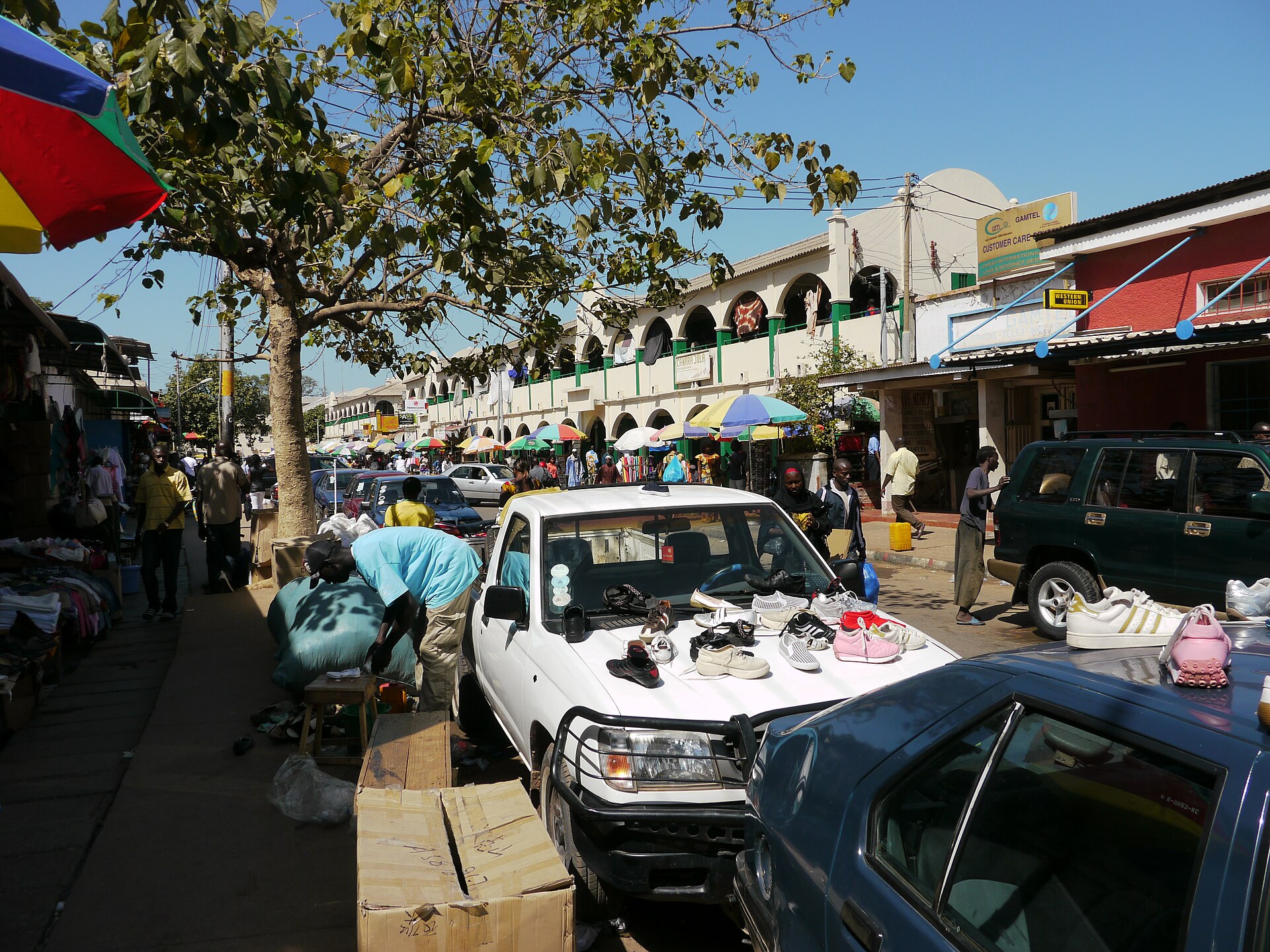 Street traders and parked cars on a busy shopping street in Banjul, with arcaded buildings in the background