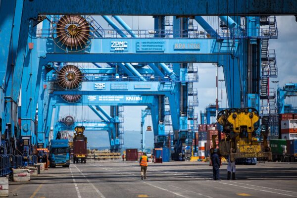Blue ship to shore gantry cranes marked KPA over a container yard at Mombasa port, with a lorry, a straddle carrier and workers on the quay