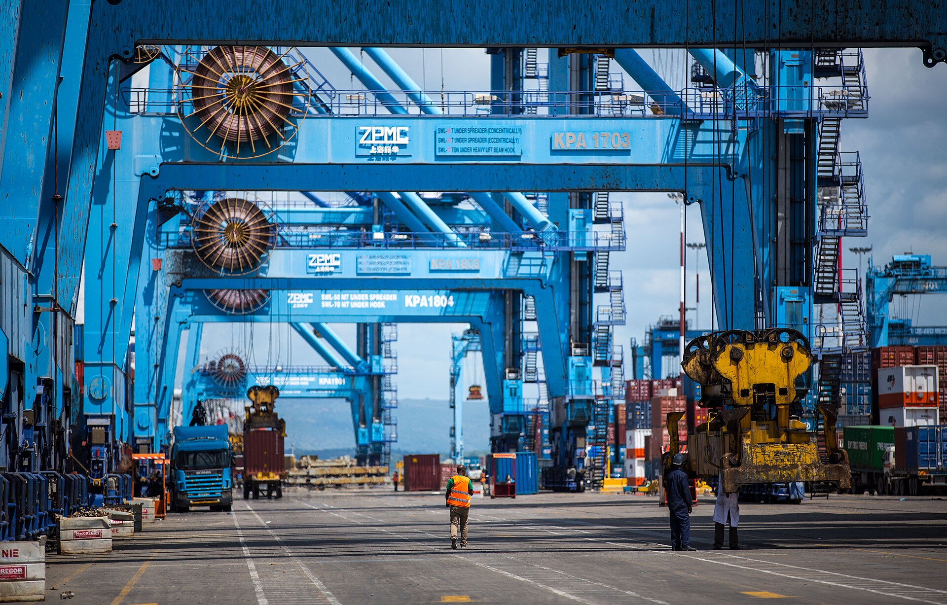 Blue ship to shore gantry cranes marked KPA over a container yard at Mombasa port, with a lorry, a straddle carrier and workers on the quay
