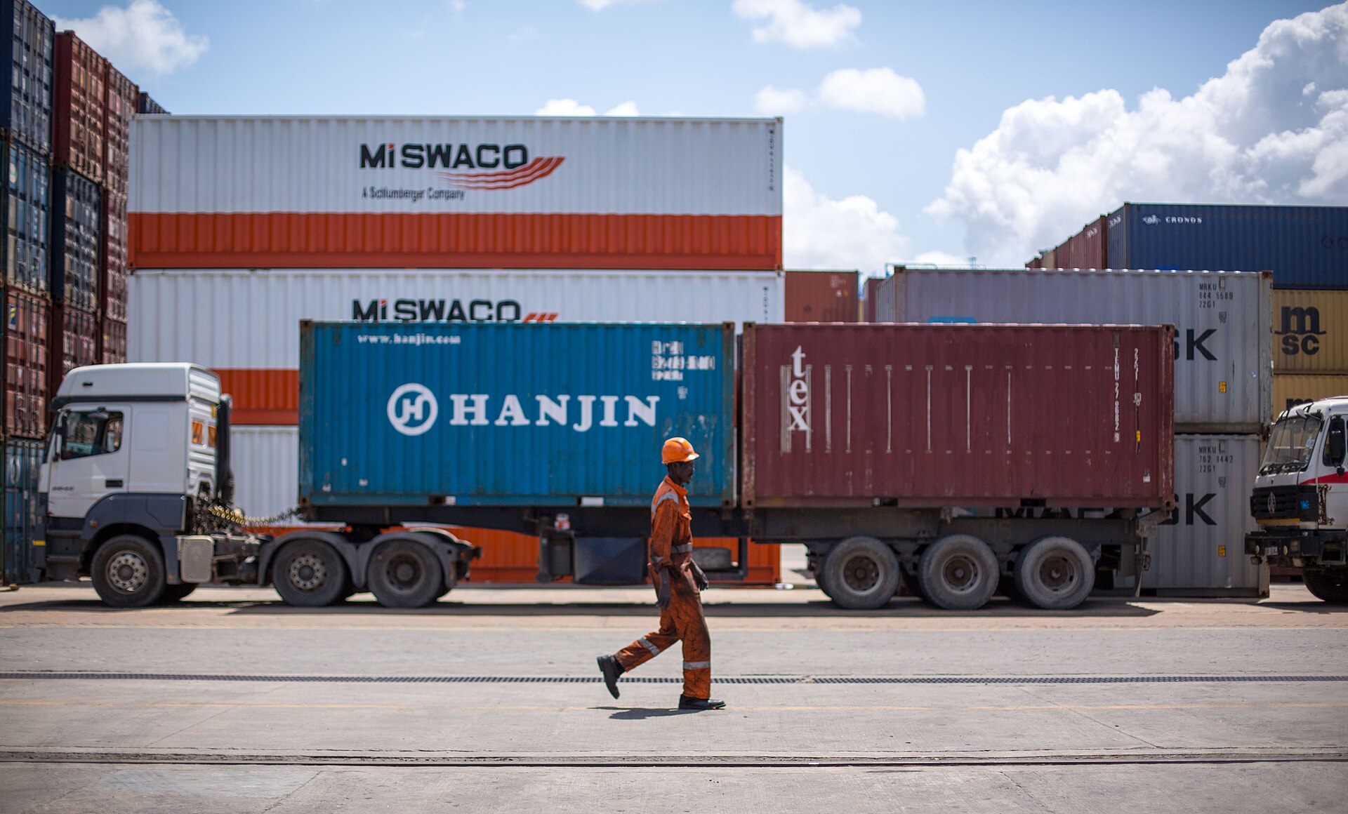 A worker in orange overalls and a hard hat walks past lorries carrying shipping containers, with stacked containers behind, at Mombasa port