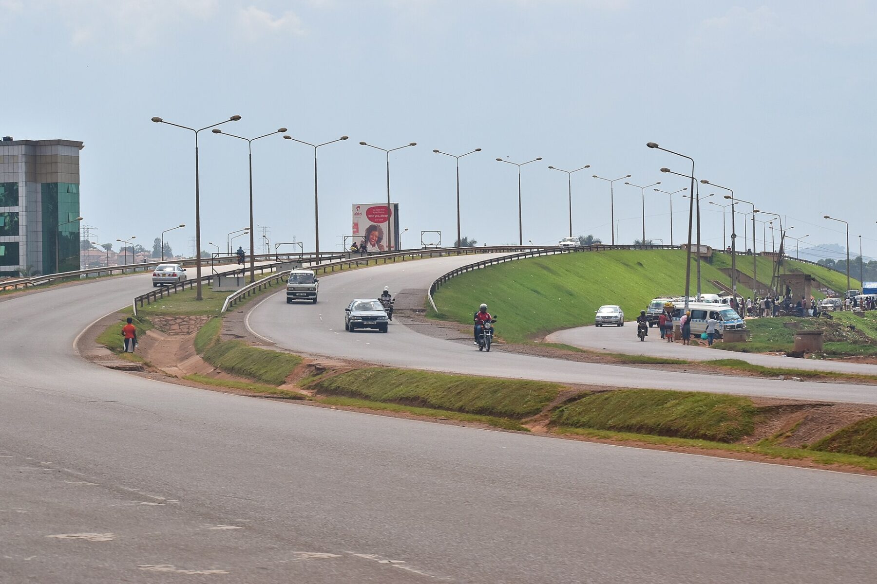 Cars and motorcycles on a raised curving road section with street lamps in the Kampala area, with a green embankment beside it