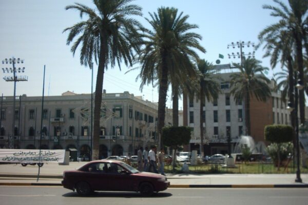 A palm-lined square in central Tripoli with a maroon car in the foreground and mid-rise buildings behind