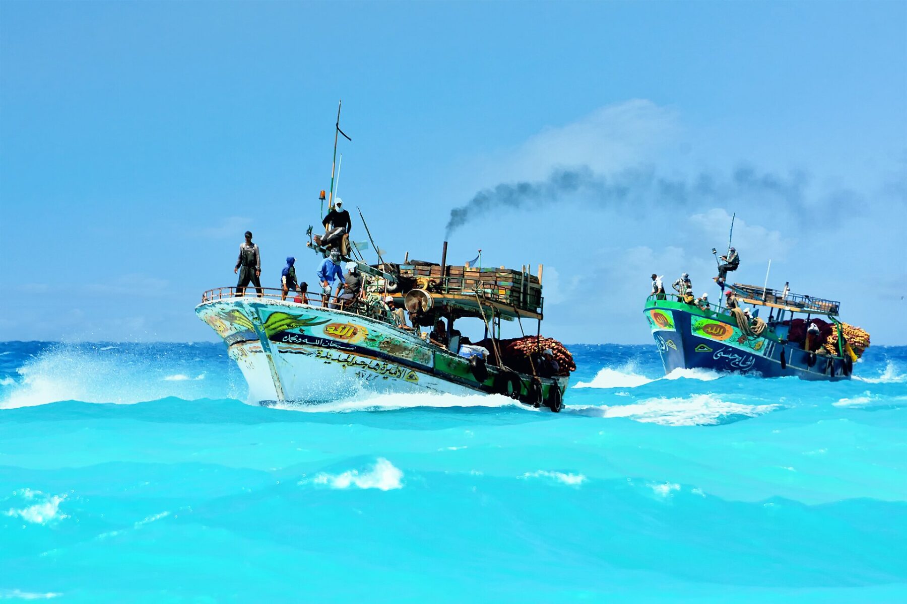 Two painted wooden fishing boats with crews on deck ride turquoise waves under a blue sky, with Arabic lettering on the hulls