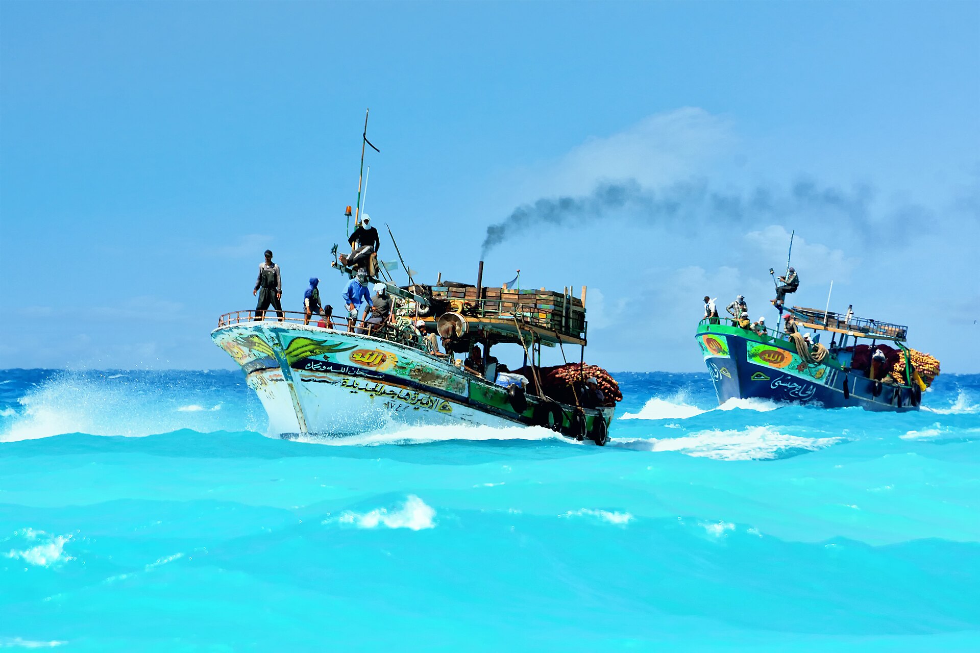 Two painted wooden fishing boats with crews on deck ride turquoise waves under a blue sky, with Arabic lettering on the hulls