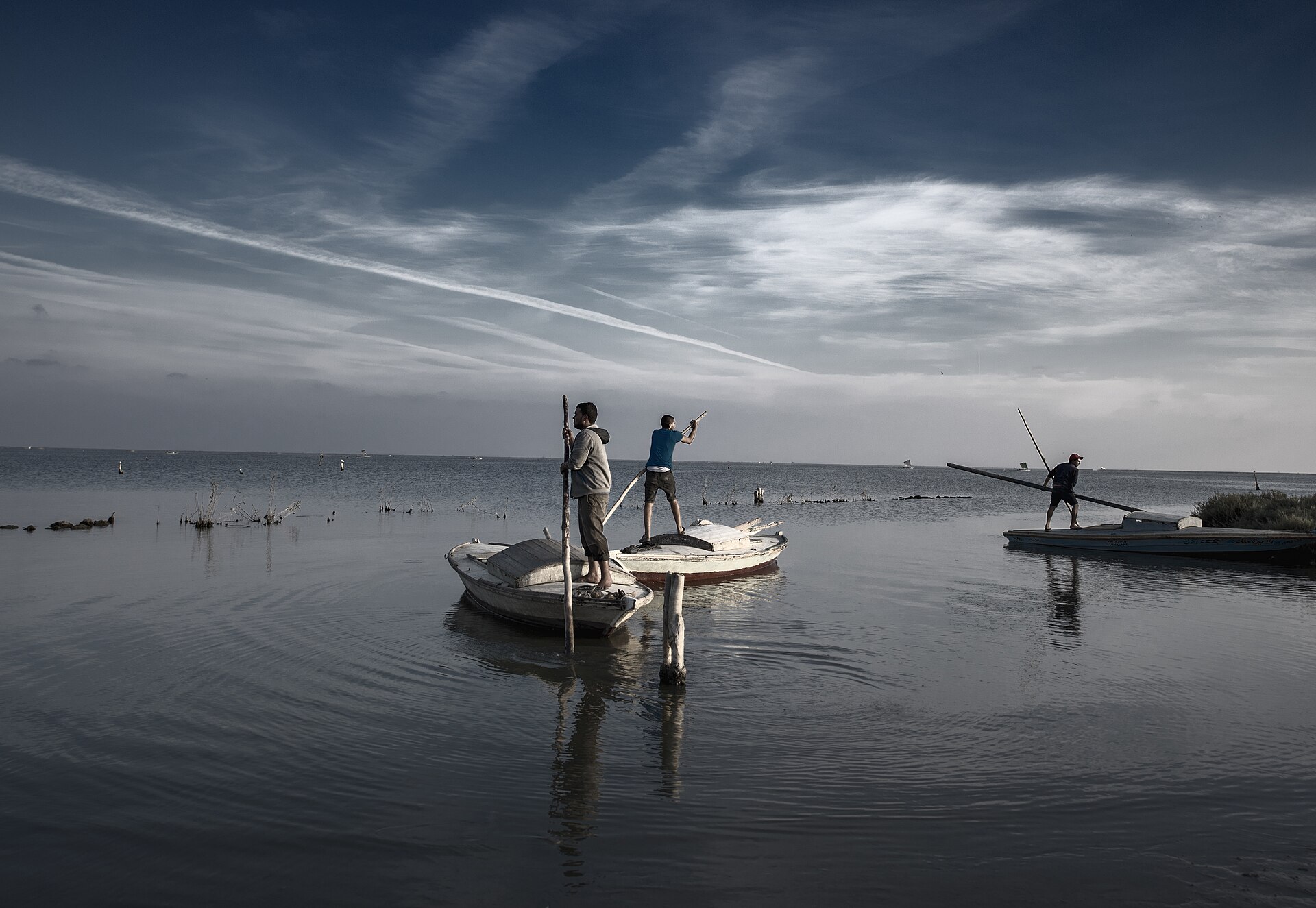 Fishermen stand in small boats on calm water under a wide sky, holding long poles, in Egypt
