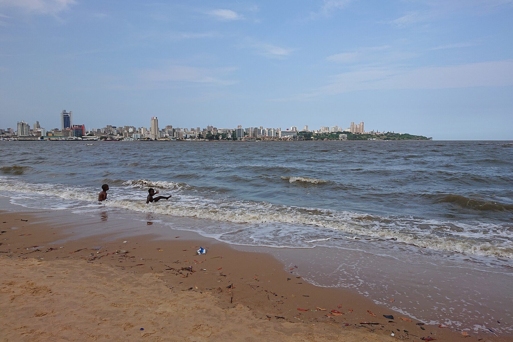 Waves reach a sandy beach in Katembe with two children in the water and the Maputo skyline on the far shore under a blue sky