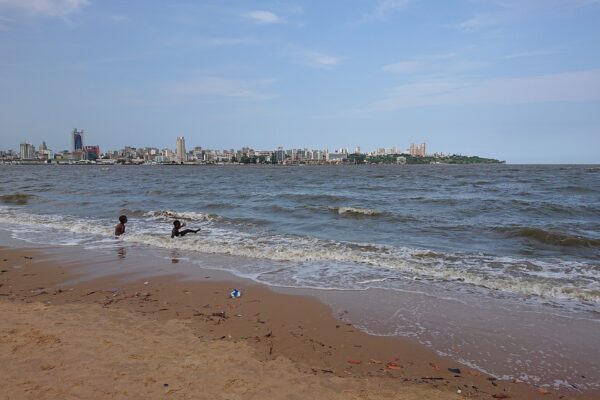 Waves reach a sandy beach in Katembe with two children in the water and the Maputo skyline on the far shore under a blue sky