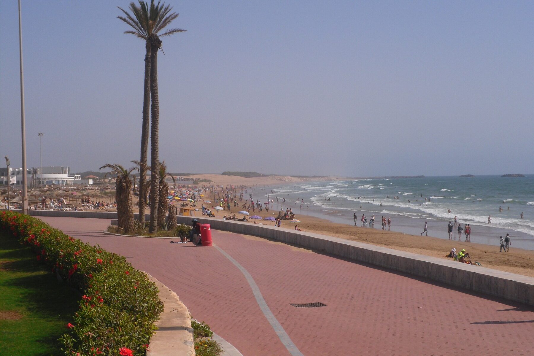 A red-brick beach promenade at Agadir with palm trees, flowering hedges and swimmers on the Atlantic beach under a hazy sky