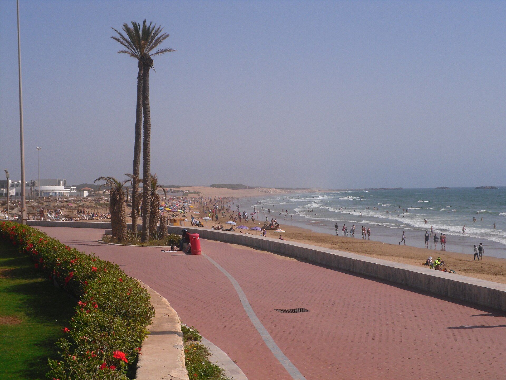 A red-brick beach promenade at Agadir with palm trees, flowering hedges and swimmers on the Atlantic beach under a hazy sky