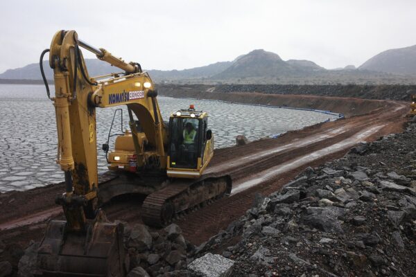 A yellow excavator on a muddy embankment beside a tailings dam, with rocky hills in the background