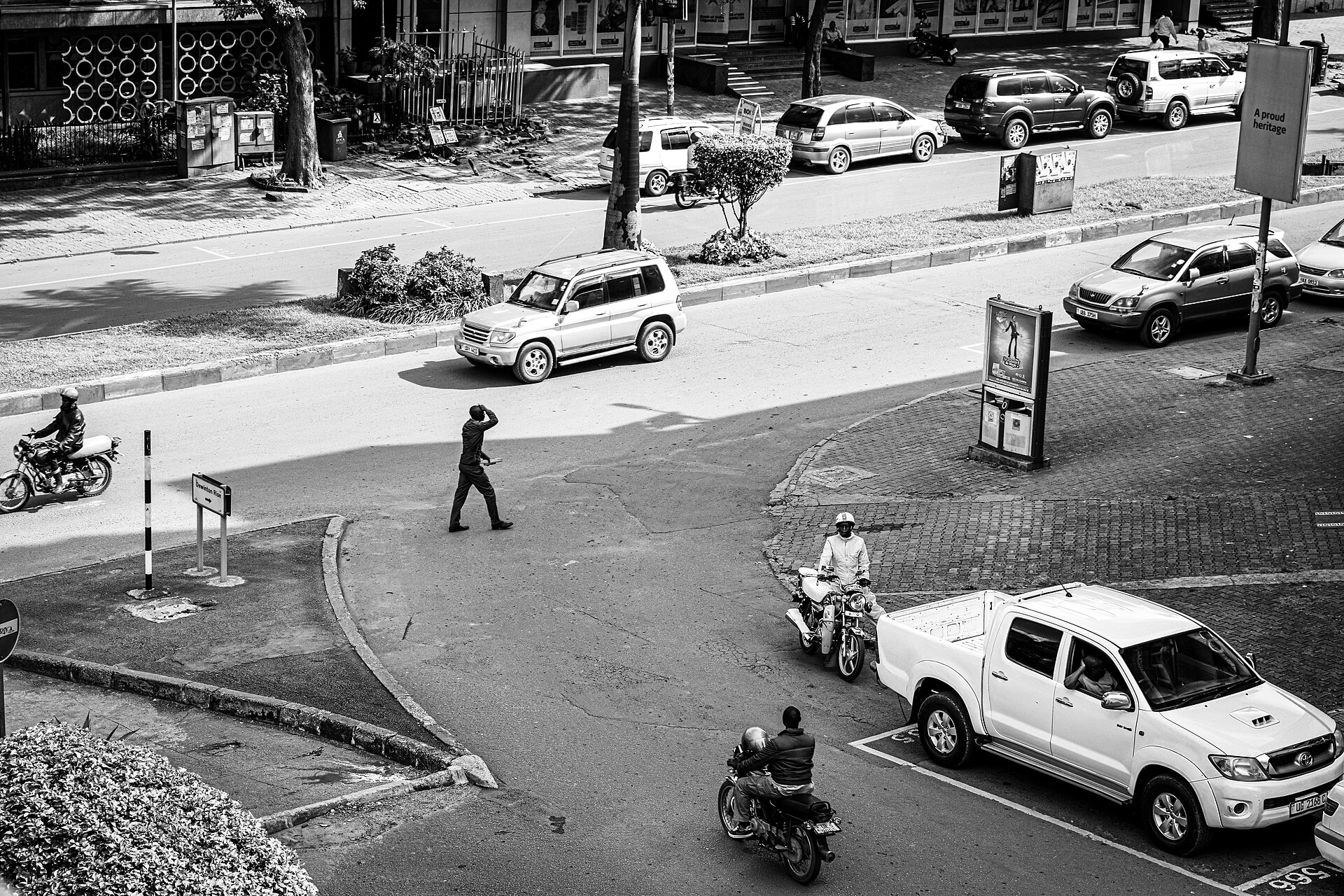 Black-and-white view from above of a Kampala street junction with cars, a white pickup, motorcyclists and a pedestrian crossing the road