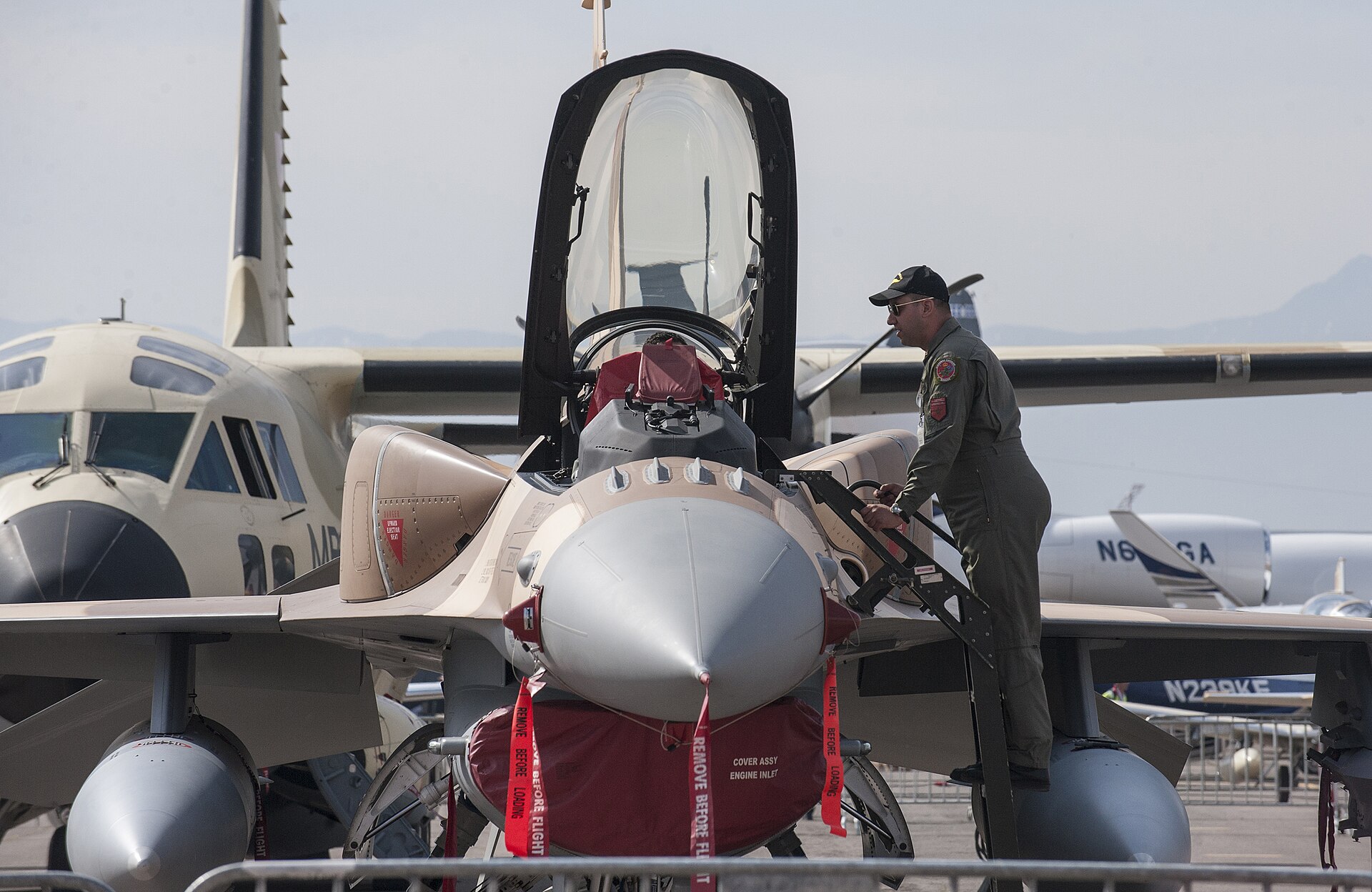 A pilot in a flight suit stands on the boarding ladder of a desert-camouflaged Moroccan F-16 with its canopy raised at an airshow