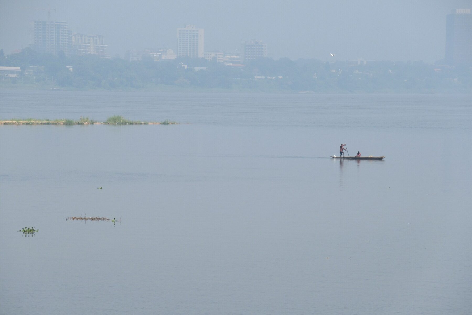A man paddles a long canoe with a passenger on the wide, hazy Congo River, with the buildings of Kinshasa and trees on the far bank