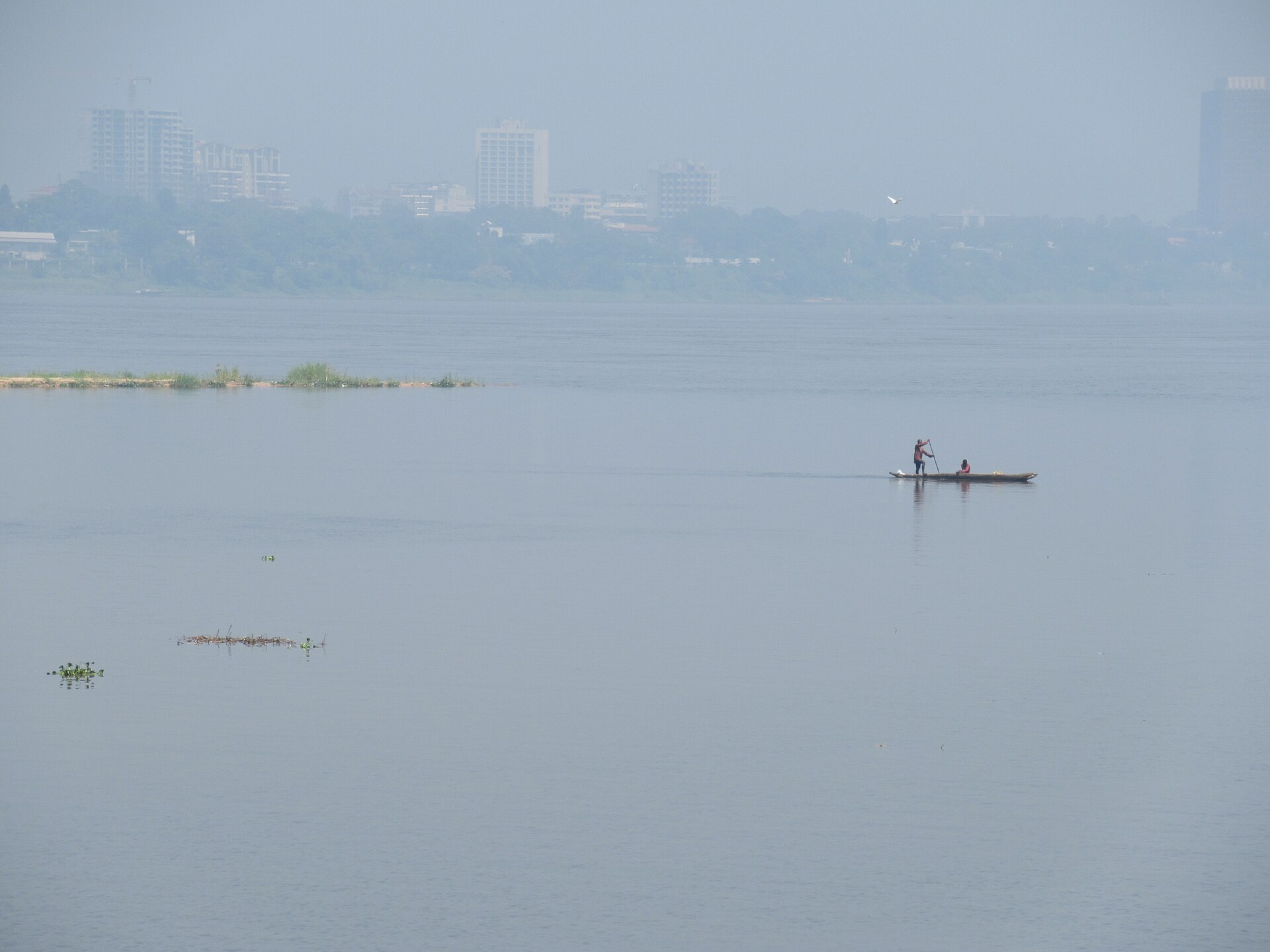 A man paddles a long canoe with a passenger on the wide, hazy Congo River, with the buildings of Kinshasa and trees on the far bank
