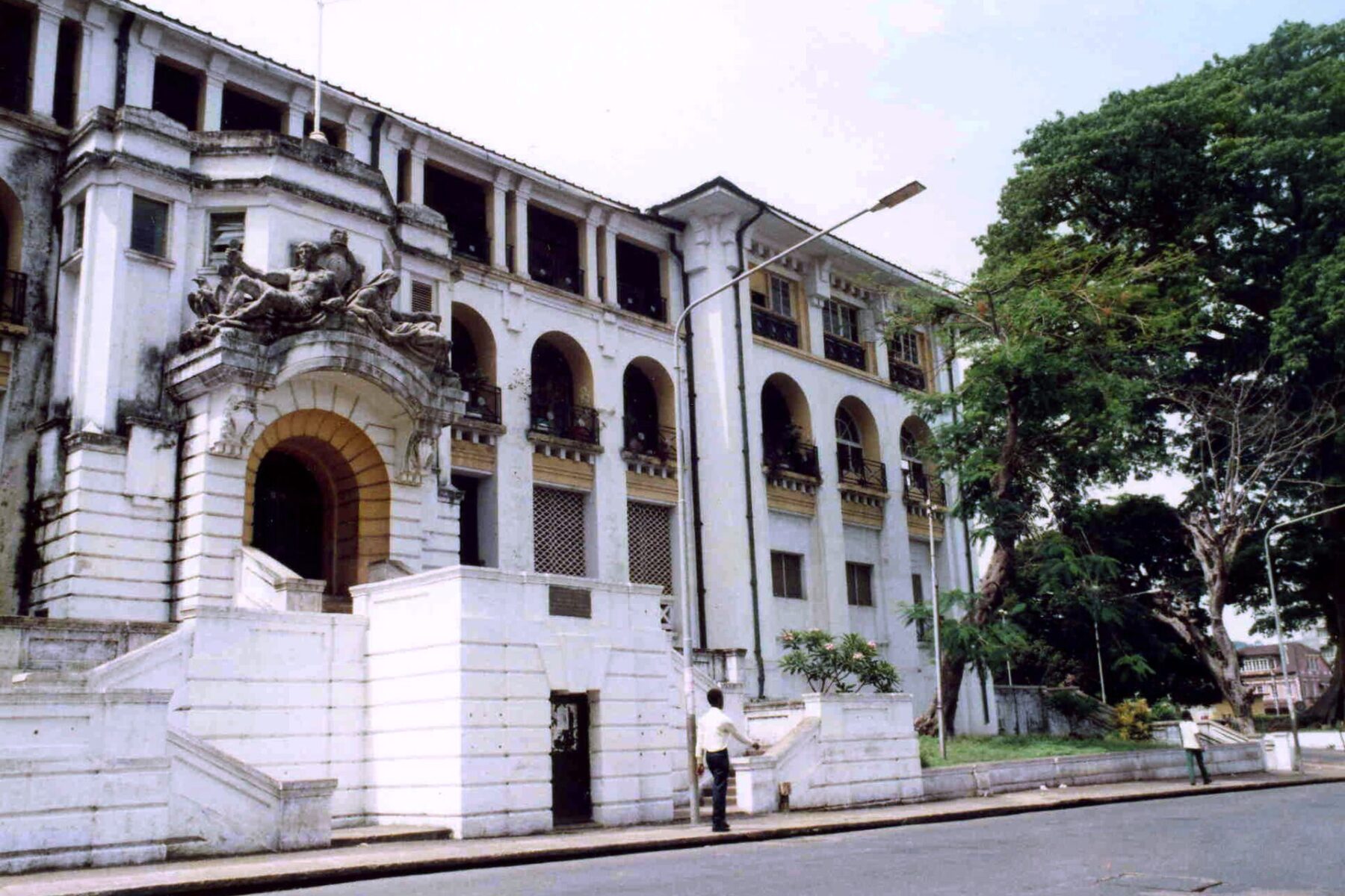 White colonial courts building in central Freetown with a carved stone group above the arched entrance and a large tree to the right