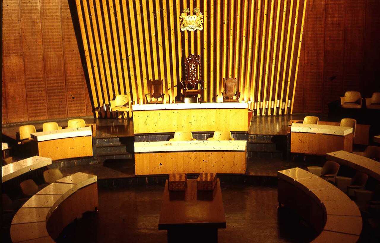 Empty wood-panelled parliamentary chamber with a carved Speaker's chair beneath a coat of arms and curved rows of desks